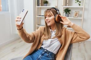 alegre jovem Loiras senhora dentro caloroso suéter dentro fones de ouvido ouço fav canções fazendo selfie vídeo ligar sentado dentro poltrona às moderno casa interior. música Tempo relaxante legal lista de reprodução conceito. cópia de espaço foto