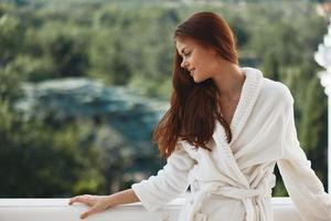 bonita mulher grandes cabelo dentro uma branco roupão de banho ficando em a sacada dentro uma hotel relaxamento conceito foto
