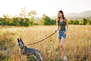 mulher caminhando dela rouco cachorro e sorridente alegremente com dentes em uma natureza andar em a Relva dentro a outono pôr do sol, estilo de vida cachorro amigo foto