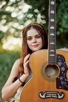 jovem hippie mulher com eco imagem sorridente e olhando para dentro a Câmera com guitarra dentro mão dentro natureza em uma viagem foto