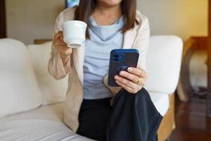 ásia mulher sentado em sofá bebericando café e assistindo conectados notícia em Móvel telefone dentro a manhã antes indo para trabalhar. suave e seletivo foco. foto
