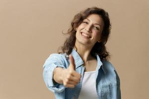 feliz sorridente encaracolado lindo fêmea dentro jeans casual camisa mostrar polegar acima gesto gostar às Câmera posando isolado em sobre bege pastel fundo. pessoas estilo de vida emoções conceito. cópia de espaço foto