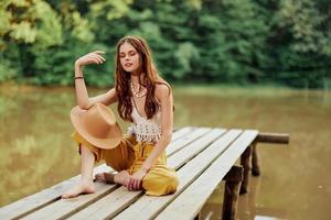 uma jovem hippie mulher senta em uma lago ponte vestindo à moda eco roupas e sorridente foto
