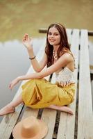 uma hippie mulher senta com dela chapéu em uma ponte de uma lago com dela mãos acima dentro a ar em uma natureza viagem e sorridente alegremente dentro roupas ecológicas. relaxado estilo de vida foto