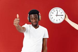 uma homem do africano aparência dentro uma branco camiseta com fones de ouvido gestos com dele mão Próximo para a relógio foto