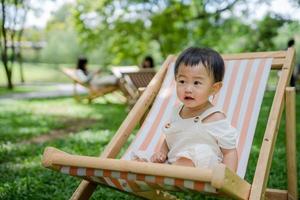 crianças ásia menina é sentado em cadeira em a grama. brilhantemente sorridente filha relaxar ou lazer com dela família em período de férias. turismo viagem com verão cafés. felicidade, risada, infância Aprendendo foto