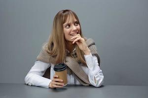 sorridente sonhando jovem Loiras aluna senhora trabalhador autonomo dentro caloroso suéter com Leve embora café reclinar em mão parece acima dentro cinzento moderno casa escritório. café pausa amante conceito. cópia de espaço. legal oferta foto