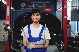 retrato do sorridente ásia masculino reparador segurando de pernas cruzadas chave inglesa às garagem profissional reparar e manutenção. carro inspeção serviço foto