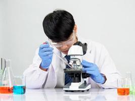 ásia masculino aluna cientista vestindo uma médico vestido dentro a laboratório olhando mão às químico. causou de mistura reagentes dentro científico pesquisa laboratórios com teste tubos e microscópio em a mesa foto