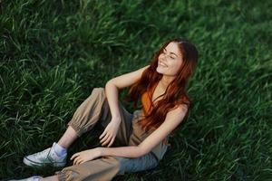 feliz mulher sorridente belas e olhando acima às a Câmera sentado em fresco verde Relva dentro a verão brilho do sol foto