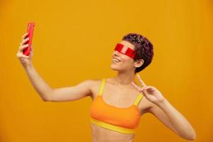 mulher blogueiro sorridente dentro incomum milenar óculos levando selfies em dela telefone dentro roupa de esporte contra a laranja estúdio pano de fundo, livre espaço foto