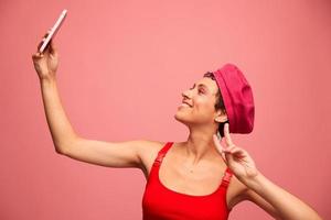 uma jovem mulher blogueiro com colori Rosa cabelo e uma curto corte de cabelo leva uma cenário do ela mesma em a telefone e transmissões uma sorrir dentro à moda roupas e uma chapéu em uma Rosa fundo monocromático estilo foto