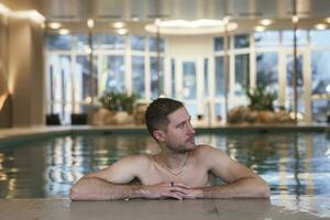 sem camisa homem olhando longe enquanto inclinado às à beira da piscina dentro luxo hotel foto