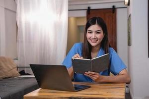 ásia mulher com uma lindo sorrir e face senta confortavelmente em dela escrivaninha às casa com uma computador portátil e caderno para relaxante e relaxante dentro uma brilhante manhã. feriado período de férias conceito. foto