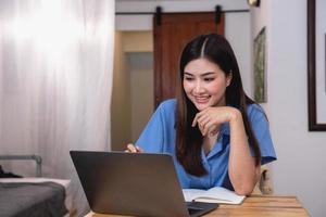 ásia mulher com uma lindo sorrir e face senta confortavelmente em dela escrivaninha às casa com uma computador portátil e caderno para relaxante e relaxante dentro uma brilhante manhã. feriado período de férias conceito. foto