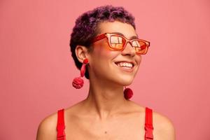 moda retrato do uma mulher com uma curto corte de cabelo dentro colori oculos de sol com incomum acessórios com brincos sorridente em uma Rosa brilhante fundo foto