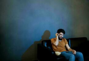 solitário homem sentado em sofá com Pipoca pratos cinzento parede foto