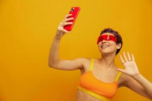 mulher blogueiro sorridente dentro incomum milenar óculos levando selfies em dela telefone dentro roupa de esporte contra a laranja estúdio pano de fundo, livre espaço foto