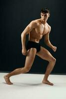 desportivo homem com uma bombeado acima muscular corpo dentro Preto calcinhas posando contra uma Sombrio fundo foto