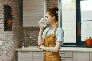 alegre mulher bebendo água dentro uma Castanho avental cozinha interior casa vida foto