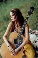 uma menina dentro roupas ecológicas hippie sentado com uma guitarra e olhando às uma pôr do sol dentro a verão foto