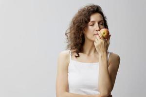 feliz encaracolado lindo mulher dentro básico branco camiseta cheirando a maçã curtiu cheiro do fruta posando isolado em sobre branco fundo. natural ecológico produtos conceito. cópia de espaço foto