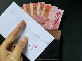 homem segurando uma branco envelope escrito do thr e Novo indonésio notas de banco, geralmente Tunjangan hari raya ou chamado thr estão dado para empregados adiante do eid. topo Visão foto