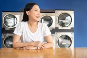 feliz ásia mulher esperando para roupas dentro uma self-service lavanderia instalação com de várias automático lavando máquinas. foto