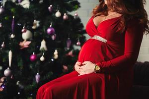 linda jovem grávida feliz em um longo vestido vermelho sentada perto de uma árvore de Natal foto