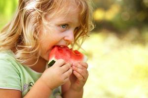 fofa pequeno menina comendo uma peça do Melancia fechar dentro a jardim dentro horário de verão foto