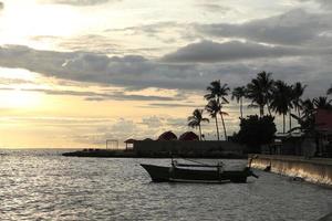 silhueta do uma de pescador barco e uma coco árvore em a de praia com uma pôr do sol fundo foto