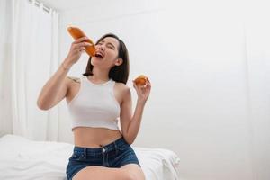 retrato do branco ásia mulher com tatuagem sentado em cama segurando pão e comendo lixo Comida. sem levando Cuidado do saúde até isto faz você gordo. gordo conceito. foto