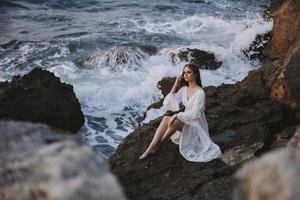 bonita mulher com molhado cabelo dentro uma branco vestir senta em uma penhasco natureza inalterado foto