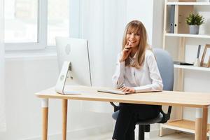 feliz sorridente adorável Loiras empresária trabalhador trabalhador autonomo Veja às Câmera sentado às local de trabalho dentro luz moderno escritório. alegre empregado trabalhos em computador conectados dentro Apoio, suporte serviço. cópia de espaço foto