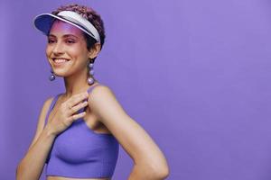 retrato do uma desportivo moda mulher posando sorridente dentro uma roxa Esportes terno para ioga e uma transparente boné em uma roxa fundo monocromático foto