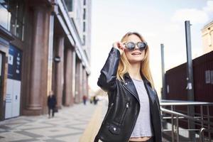 elegante mulher dentro uma couro Jaqueta e oculos de sol perto a construção foto