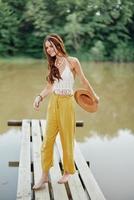 uma jovem mulher dentro uma hippie Veja e vestido ecológico dançando ao ar livre de a lago vestindo uma chapéu e amarelo calça dentro a verão pôr do sol foto