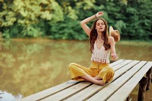 uma jovem hippie mulher senta em uma lago ponte vestindo à moda eco roupas e sorridente foto