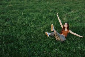 uma jovem mulher jogando jogos dentro a parque em a verde Relva espalhando dela braços e pernas dentro diferente instruções queda e sorridente dentro a verão luz solar foto