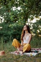 uma jovem hippie mulher medita dentro natureza dentro a parque, sentado dentro uma lótus posição em dela colorida xadrez e desfrutando harmonia com a mundo dentro roupas ecológicas dentro a outono foto