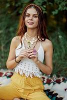 jovem hippie mulher sentado dentro natureza em uma xadrez com uma flor dentro dela mãos e olhando às a Câmera sorridente Atenciosamente dentro roupas ecológicas dentro outono dentro a pôr do sol foto