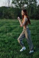 uma jovem mulher exercício lado de fora e corrida dentro a parque. dentro a tarde em a verde Relva para dentro a pôr do sol foto