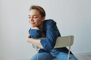 uma jovem mulher sentado dentro uma cadeira às casa sorridente com dentes com uma curto corte de cabelo dentro jeans e uma jeans camisa em uma branco fundo. menina natural poses com não filtros foto