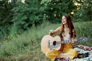 jovem fêmea hippie artista tocam guitarra e canta canções dentro ecológico roupas sentado em a terra lado de fora dentro natureza dentro a outono olhando Fora às a pôr do sol foto