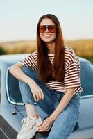 uma elegante mulher sorrisos docemente dentro à moda oculos de sol, uma listrado camiseta e jeans, senta em a tronco do uma carro e parece às a lindo natureza do outono. viagem estilo de vida foto