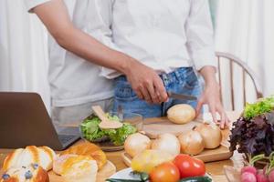 jovem ásia casal cozinhando com frutas e legumes e usando computador portátil dentro a cozinha para cozinhar Comida juntos dentro a família Felizmente, família conceito. foto