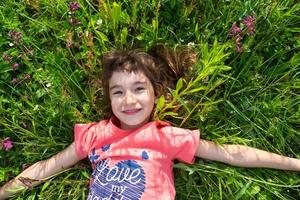 retrato do uma criança menina dentro verão deitado dentro a Relva e flores silvestres com calcanhares e Palmeiras. verão tempo, liberdade foto