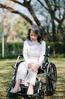 ásia velho mulher sentado em uma cadeira de rodas ao ar livre dentro a parque ter dor dentro a braços, pulsos e corpo dentro Sol luz foto