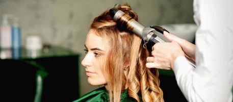 cabeleireiro usando ondulação pinças cachos grandes Castanho cabelo em a jovem caucasiano menina dentro uma beleza salão. foto