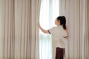 jovem empregada abrindo cortinas em quarto de hotel